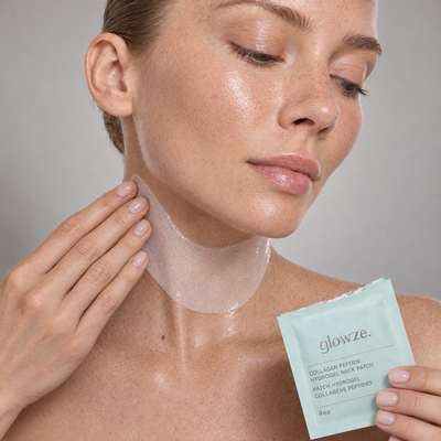 Freckled woman applying a glowze. hydrogel neck patch, holding the mint glowze. sachet, grey studio backdrop